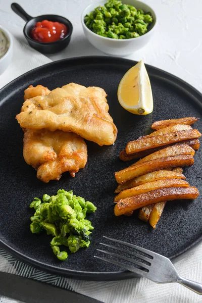 British Traditional Fish and chips with minty mashed peas detail and a slice of lemon. on black round plate over white lintn and stone surface