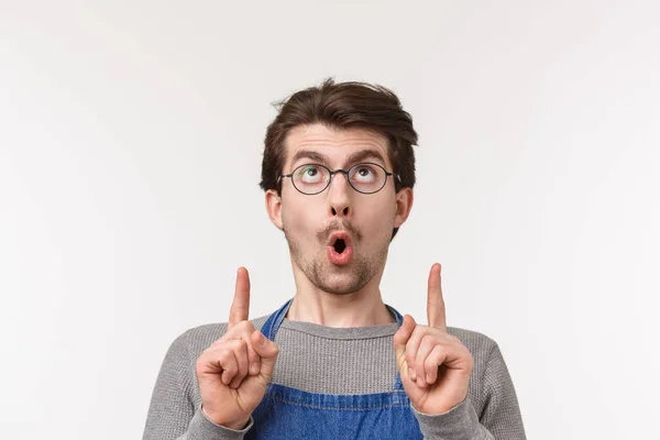 stock image Close-up portrait of interested and excited caucasian bearded guy in apron and glasses, say wow and stare pointing fingers up at something cool, spot awesome product, white background