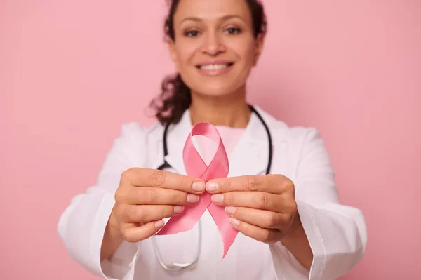 Soft focus on Breast Cancer Awareness Pink Ribbon, in hands of blurred mixed race female doctor in white medical coat, isolated on colored background. 1 st October, World Day of fight breast cancer