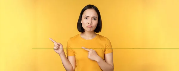 不安的亚洲女人指着左边的手指 显得有些失望 抱怨横幅 站在黄色的背景上 — 图库照片