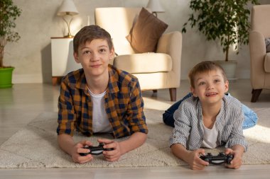İki neşeli çocuk bir odada yere uzanıp joysticklerle video oyunları oynuyor.