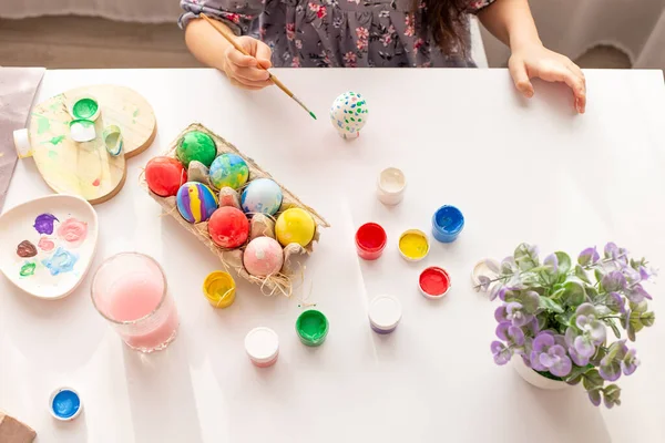 Hands of a little girl, with a painted egg, on a white table with multi-colored eggs in a tray, brushes and paints.