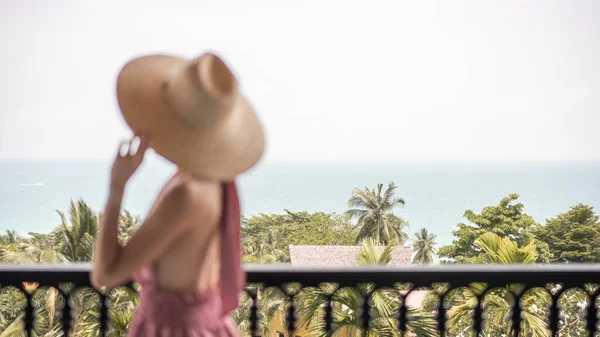 Blur side view of woman in pink dress and straw hat standing on a hotel balcony, ocean view.
