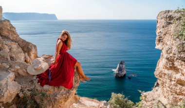 A girl with flowing hair in a long red dress sits on a rock above the sea. The stone can be seen in the sea.