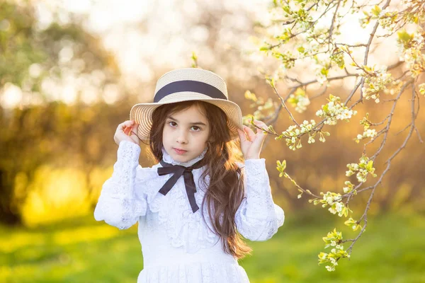 花の木の下に立って 帽子と白いドレスの美しい少女の肖像画 — ストック写真