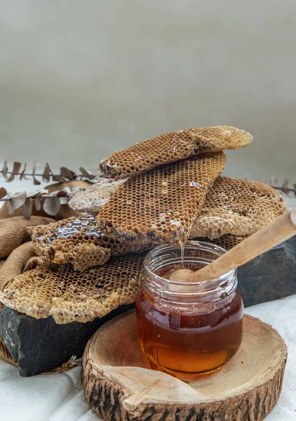 美味的蜂蜜从新鲜的蜂窝滴在玻璃瓶上 用木制蜂蜜搅拌器粘在黑色的岩石上 — 图库照片