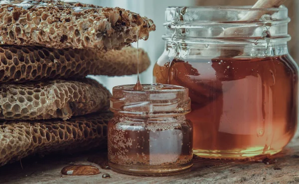 将新鲜蜂窝中的美味蜂蜜滴在玻璃瓶上 用木制蜂蜜搅拌器棒粘在旧木桌上的特写 — 图库照片