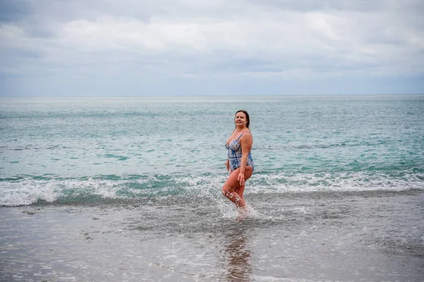 水着姿の豊満な女がサーフィン中に水の中に入る ビーチで一人で 雲の中に灰色の空 冬に泳いで — ストック写真