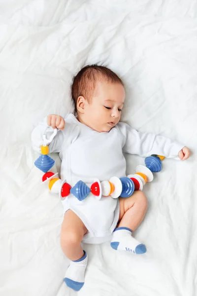 Baby boy holds colorful rattle toy. Top view of little kid lying on crumpled white linen with his first toy. Newborn in bed.