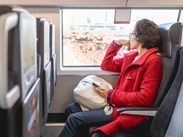 赤いダッフルコートを着た笑顔の女性が電車の郊外の窓の近くに座っています 陸路車両での移動 — ストック写真