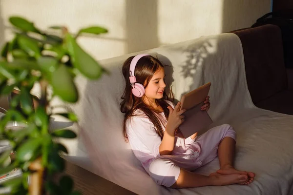 Little Adorable Girl Pink Headphones Sits Couch Looks Digital Tablet — Zdjęcie stockowe