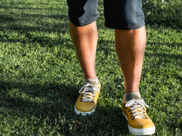 Man in bright yellow sneakers is standing on green grass lawn in park. Modern hipster\'s shoes. Urban fashion.