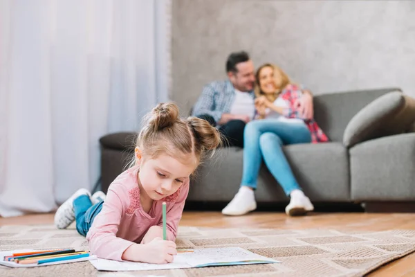 Маленькая Девочка Рисует Книгу Лежащую Полу Пока Любящие Родители Сидят — стоковое фото