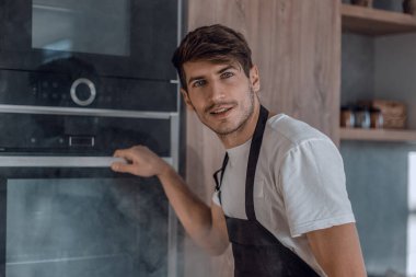 Genç adam ev mutfağında kırık fırının yanında duruyor.