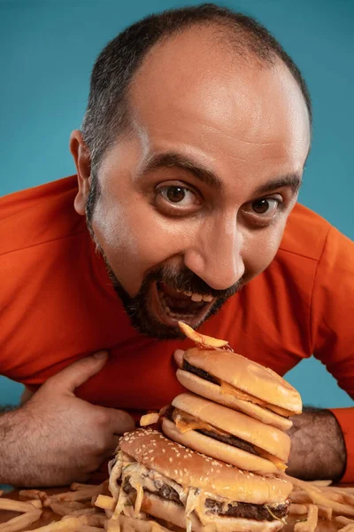髭を生やした中年男性が赤いタートルネックに身を包み ハンバーガーとフライドポテトでポーズをとっている姿がクローズアップされています 青の背景 ファーストフード — ストック写真