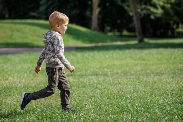 可爱的男孩笑着跑过草地 一个敏捷快乐的孩子正在一座夏季城市公园里做运动 — 图库照片