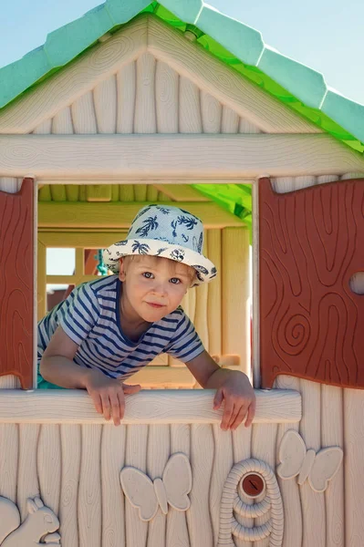 他是个戴着帽子的快乐的小男孩 在一间玩具小屋里 他用百叶窗直勾勾地看着窗外的相机 — 图库照片