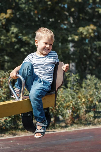 一个快乐的 微笑的小男孩在城市操场的秋千上 背靠着绿叶的背景 — 图库照片