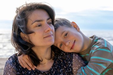 Cute happy boy hugs his mom by the sea. looking into the camera