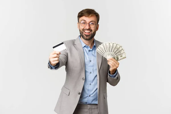グレーのスーツと眼鏡の髭のビジネスマンの肖像 クレジットカードとお金を示す 幸せな笑顔 白い背景に立って — ストック写真
