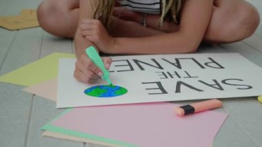 Kadın ellerinin yavaş çekimde posterler çizerek gezegeni kurtardığını gösteriyor..
