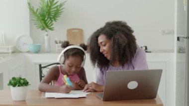 Happy multiethnic women single mom sits next adorable baby girl in wireless headphones Mom her baby girl drawing together colorful images with felt-tip pens watching online course on laptop at home