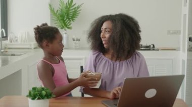 Multitasking young African mother freelance worker, entrepreneur working remotely on laptop, while her beautiful little daughter presenting her a gift for International Mothers Day