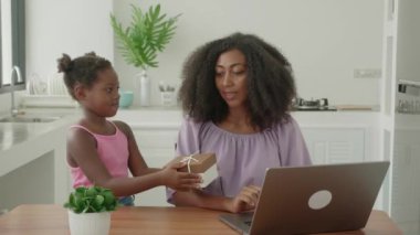 Multitasking young african american woman freelance entrepreneur working remotely on laptop while her beautiful little daughter is presenting her a birthday present. Mother and daughter hugging