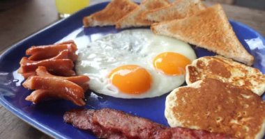 Close up fried unhealthy breakfast On big blue plate, an unhealthy breakfast lies on table at daytime Unhealthy breakfast concept. Slow motion morning menu consists of fried egg, sausage toast bacon