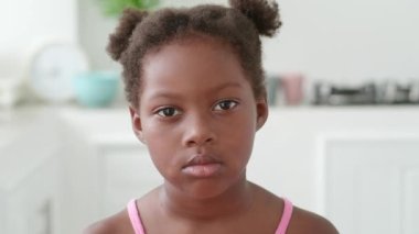 Portrait of sad kid against the backdrop of apartment. sad kid with dark-eyedstares piercingly straight into camera. sad kid female standing in bright room concept of negative emotions