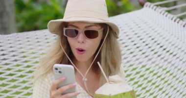 woman with glasses sits on hammock in resort and holds surprised woman on vacation says wow while flipping through the news feed on her smartphone. Internet addiction to social networks on smartphone