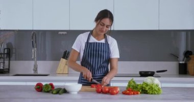Önlüklü genç ev hanımı, geniş bir mutfakta salata pişiriyor. Ev hanımı, 25-29 yaş arası sebze, domates doğrama ve salata pişiriyor. Evde salata pişirme kavramı.