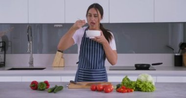 Tatsız yemek, tuzsuz salata, önlüklü ünlü modern mutfağın arka planına karşı yemek hazırlar, kadın tuzsuz salata dener ve tuzsuz salata tarifinde başarısız olur.
