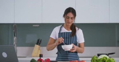 Mutfaktaki kadın salata deniyor ve tatmin olmuş bir yüz tarifi gösteriyor. Mutfakta duran önlüklü başarılı bir kadın salata yiyor ve onaylı bir şekilde tarifini onaylıyor. Lezzetli kahvaltı tarifi başarılı oldu.