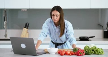 Mavi gömlekli çekici bir kadın çevrimiçi yemek pişiriyor. Video dersleri alıyor. Salatanın tutarlılığını kontrol ediyor.