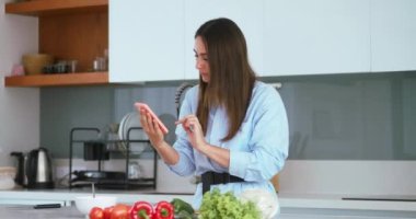 Mutlu genç bir kadın sebze yemeği hazırlıyor. Çevrimiçi vegan tarifini akıllı telefondan kontrol ediyor. Gülümseyen vejetaryen bir kız modern mutfakta mobil yemek kitabı menüsü uygulamaları kullanarak sağlıklı yemek salatası pişiriyor.