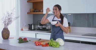 Çizgili önlüklü, genç, güzel bir kadın tatsız bir salata ve tiksinti içinde somurtmayı deniyor. Modern olmayan bir mutfakta duran bir kadın tatsız bir salata yer ve olumsuz duygular gösterir.
