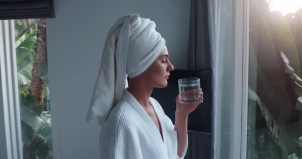 身穿白色浴衣和毛巾的妇女在透明的玻璃杯中喝水时 有着健康的习惯 站在窗边的年轻女子的健康习惯概念与黎明的侧面景色相遇 健康的习惯很神奇 — 图库视频影像