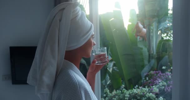 有用的习惯概念穿着白袍的女人在清澈的玻璃中喝水早起 有用的习惯现代室内环境中的女人在欣赏黎明后的景象 — 图库视频影像