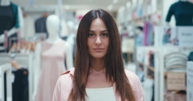 Yakın plan portre müşterisi serginin ortasında duruyor. Alışveriş ve kıyafet satışı konsepti. Genç, koyu renk saçlı kadın elbise satıldığı gün elbise askılarının yanında duruyor.