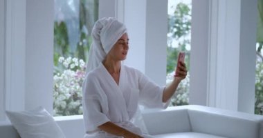 Kafasında havlu olan beyaz önlüklü kadında akıllı telefon konsepti büyüklüğünde video çağrısı var. Akıllı telefondan konuşan ve modern iç mekan görüntülerinde el sallayan kadın.