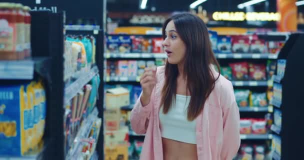 ダークヘアの顧客はスーパーマーケットのカウンターで商品を検査し 過剰に憤慨しています 若い女性は店の背景に過剰な商品に驚いています インフレーションコストから損失の女性 — ストック動画