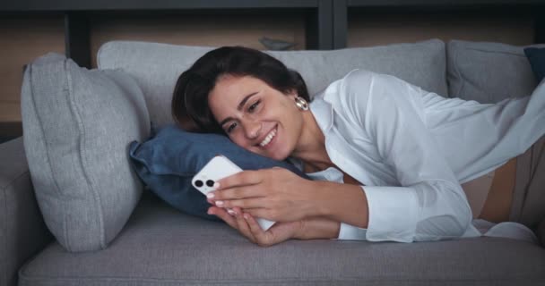 女性はソファーに横たわり スマートフォンを持っています サイトの関係構築 女性はニュースフィード 関係構築の概念をスクロールします 女性は笑顔で 異性とメッセージを交換する — ストック動画