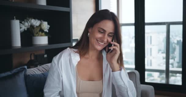 年轻的女人在用智能手机打电话 女性坐在一个可以俯瞰摩天大楼的现代化公寓里 依靠智能手机技术和创新 远程通信 透过电话分享情绪 — 图库视频影像