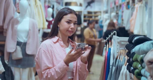 女人站在商店里打电话咨询朋友关于服装的选择 服装的选择反映了幸福的友谊服装的选择在科技的帮助下象征着现代生活方式 — 图库视频影像