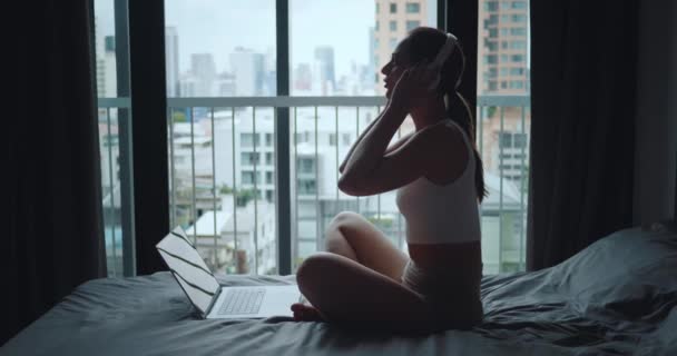 ベッドルームのベッドに座っている女性は ヘッドフォンを離れて 街の窓の景色を見ながらノートパソコンを閉じます リモートからリラクゼーションまで仕事の終わり 仕事の落ち着きのウィンドウで考える — ストック動画