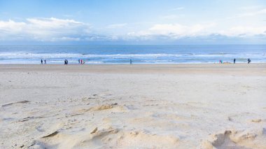 Sonbaharda bulutlu bir gökyüzünün altında gezinen birkaç kişiyle alçak gelgit sırasında geniş kumlu bir sahili gösteren sakin bir sahil sahnesi..