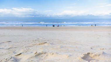 Sonbaharda bulutlu bir gökyüzünün altında gezinen birkaç kişiyle alçak gelgit sırasında geniş kumlu bir sahili gösteren sakin bir sahil sahnesi..