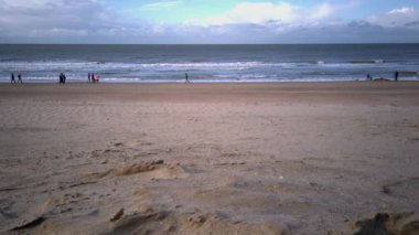 Sonbaharda bulutlu bir gökyüzünün altında gezinen birkaç kişiyle alçak gelgit sırasında geniş kumlu bir sahili gösteren sakin bir sahil sahnesi..