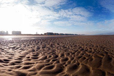 Geniş, kumlu bir sahil. Karmaşık desenleri olan kumsal, parlak mavi gökyüzünün altında, uzak bir şehrin siluetine doğru uzanıyor. Huzurlu ve davetkar sahne, kıyı manzarasının sükunetini yakalar..